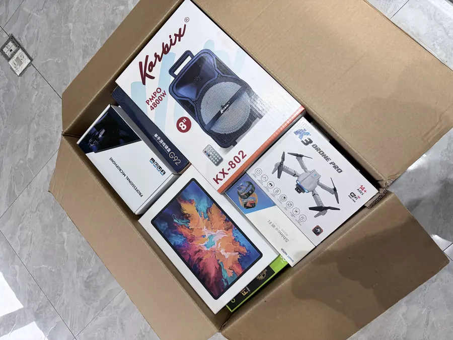 Open wholesale carton showing electronics packed as a visible top layer: portable speaker, drone, watch and accessories